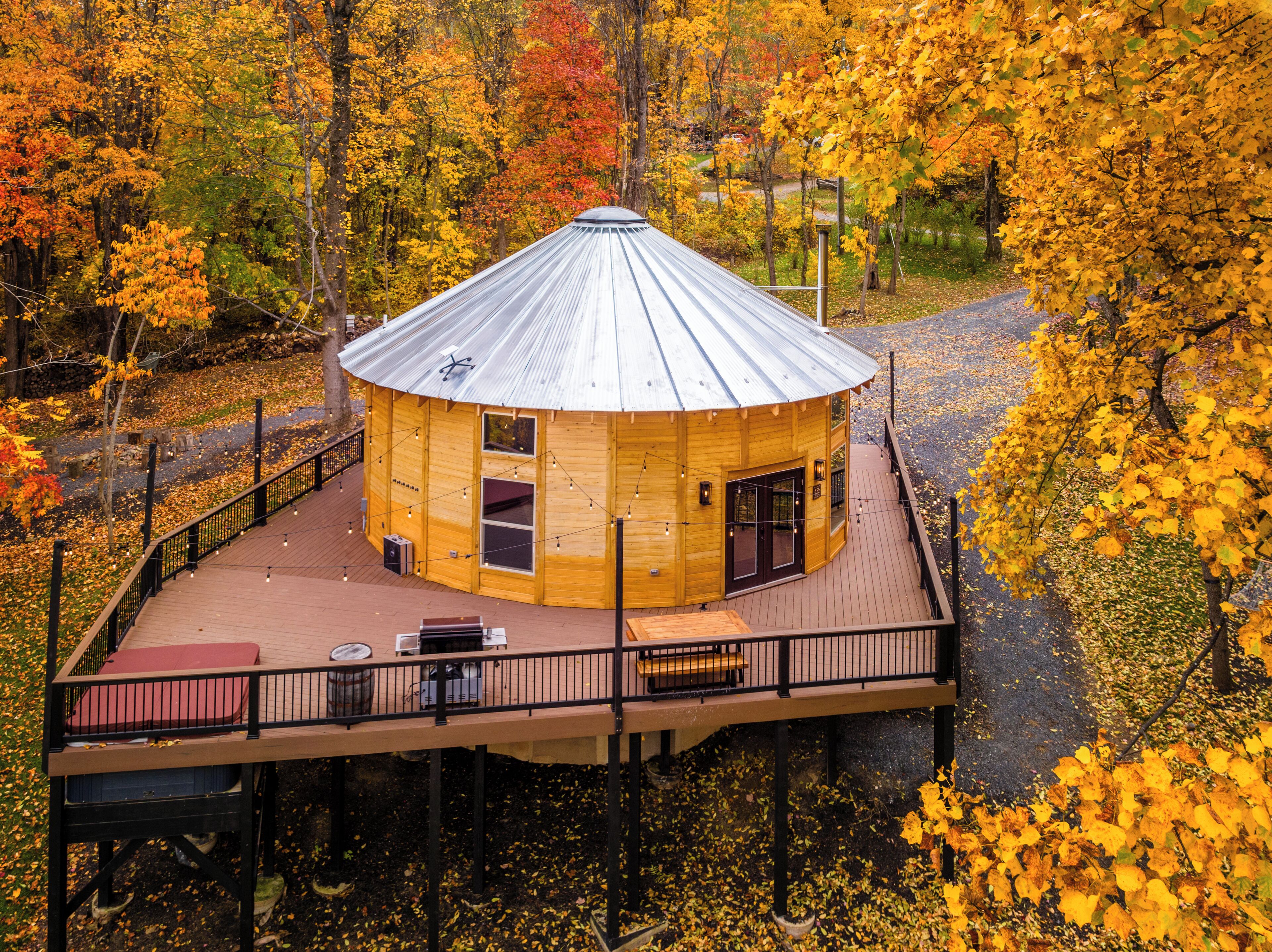 A wooden cabin with a metal roof, surrounded by a wooden deck and a railing, set in a forest with autumn foliage.