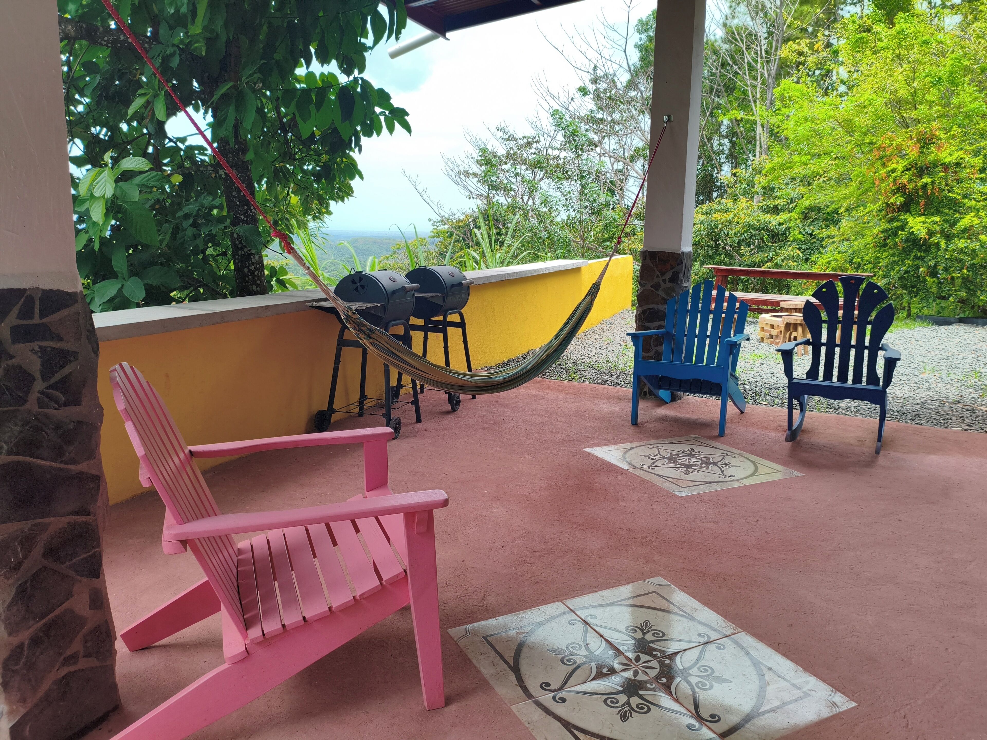 A patio with a pink chair, a blue chair, and a hammock. There are barbecue grills and a yellow wall in the background.