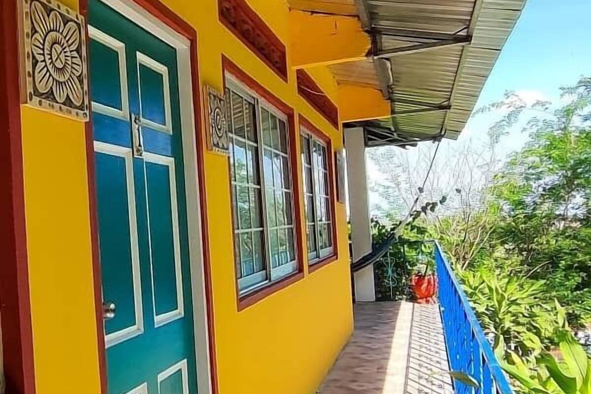 Un edificio con fachada amarilla brillante, una puerta y barandilla azules, y vistas a un jardín.