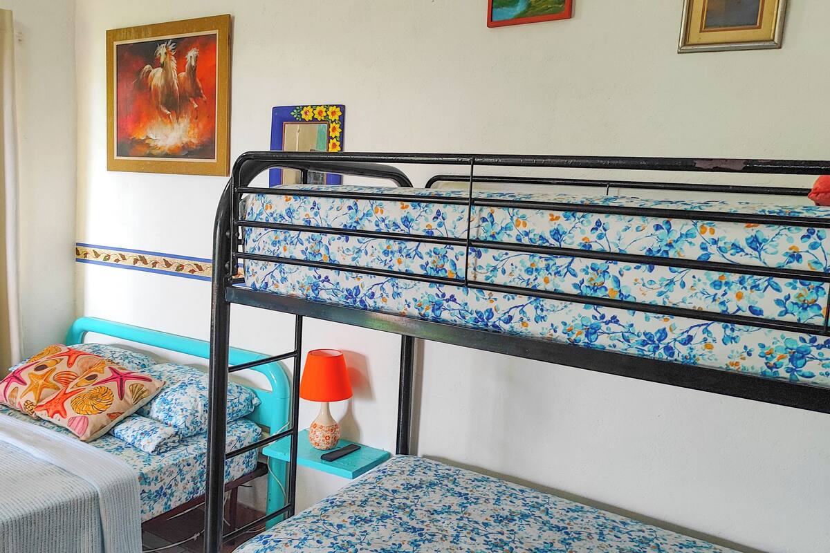 Una cama litera con colchón estampado floral, una mesita pequeña con mantel blanco y una lámpara roja sobre una mesita de noche azul.