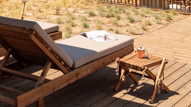 Une terrasse en bois avec un fauteuil de relaxation et une petite table, offrant une vue dégagée sur une plage, l’océan et une pinède.