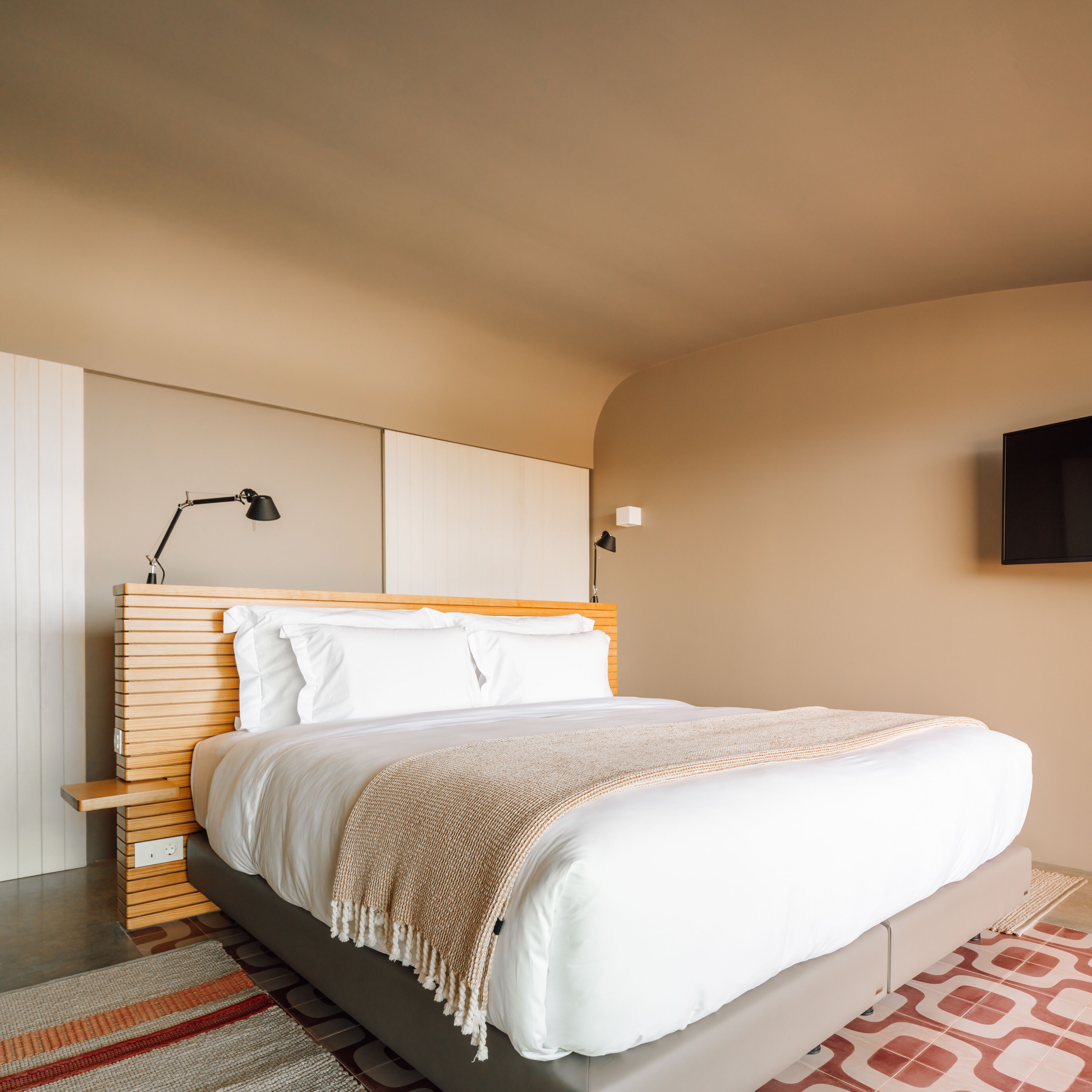 A modern hotel room with a wooden headboard, a large bed with white linens, a beige throw blanket, a bedside table, and a flat-screen TV mounted on the wall.