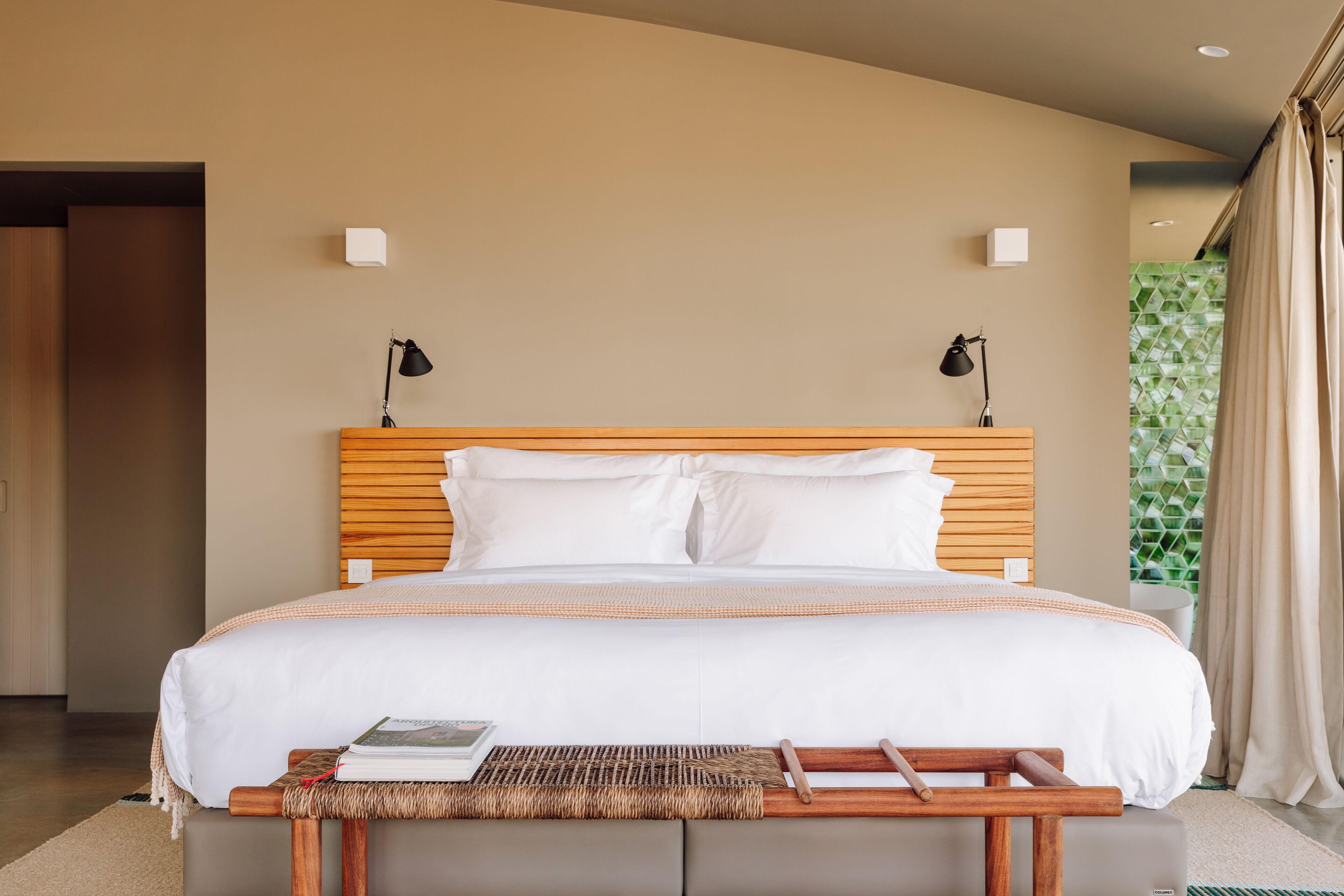 Ein modernes Hotelzimmer mit einem großen Bett, zwei Nachttischlampen, einem Holz-Kopfteil und einem kleinen Nachttisch mit einem Buch.