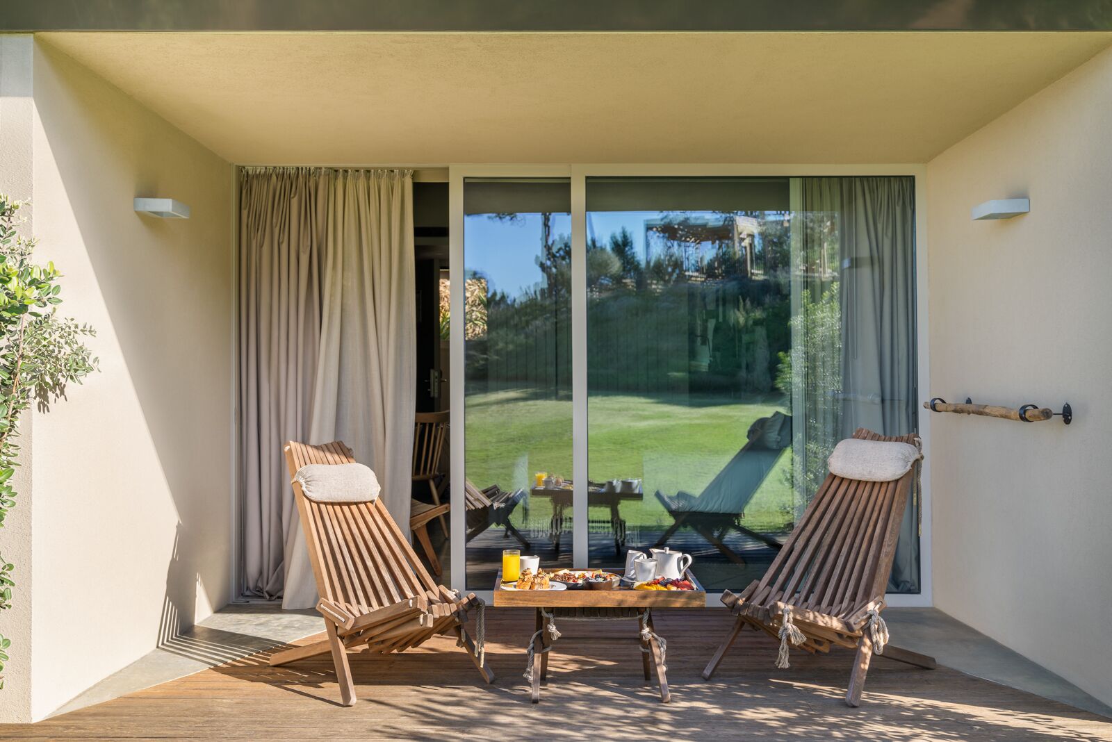 Een zonnig terras met twee houten lounge fauteuils, een tafel met ontbijtartikelen, en uitzicht op een groen gazon door grote glazen deuren.