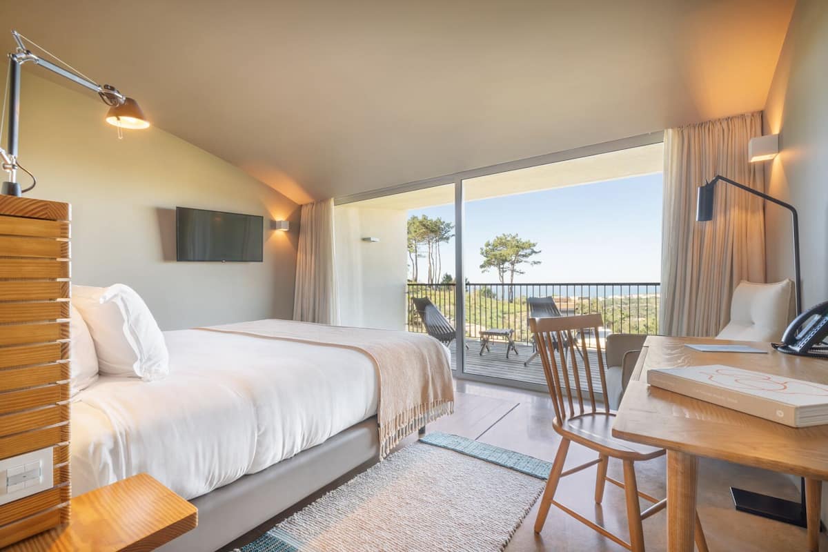 Habitación de hotel con una cama grande, un escritorio con una silla, un teléfono y un televisor. Tiene un balcón con vistas al paisaje.