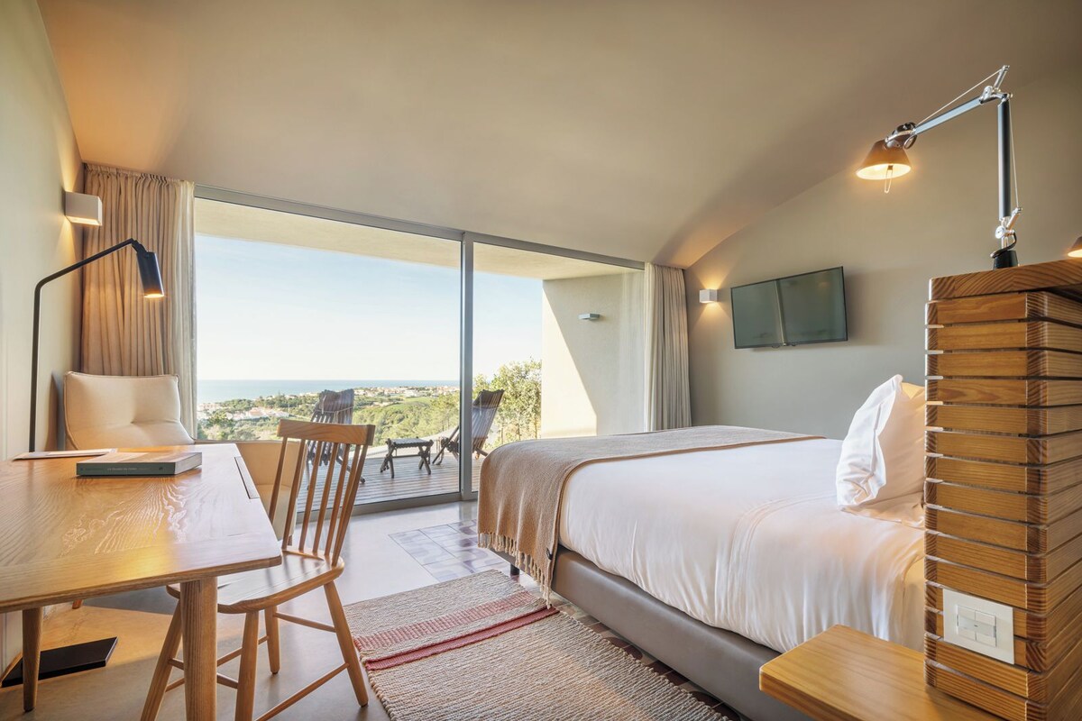 A hotel room with a bed, a desk, and a chair. There is a view of the outdoors through a large window.