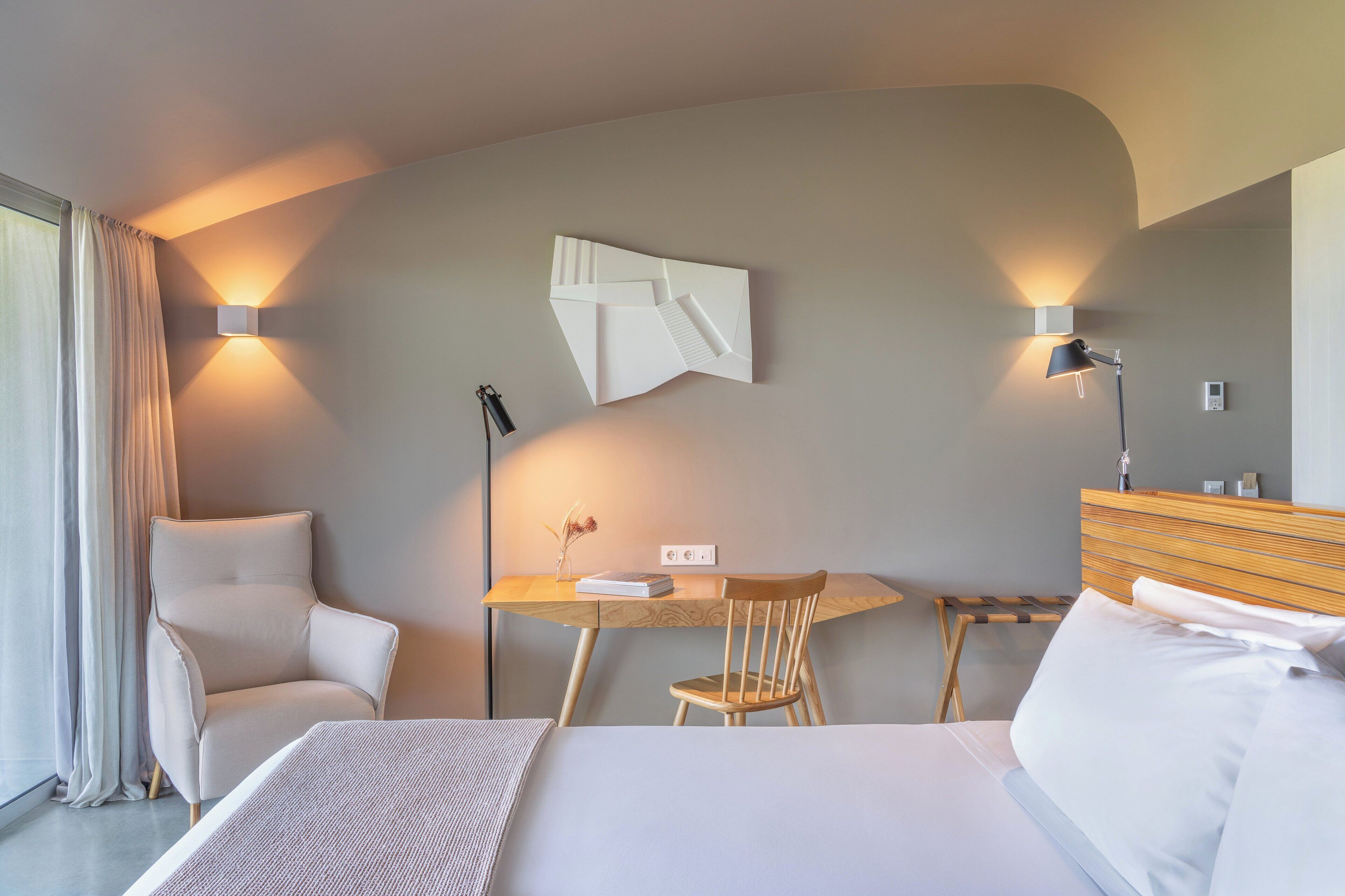 A modern hotel room with a bed, a desk, a chair, and a lamp.