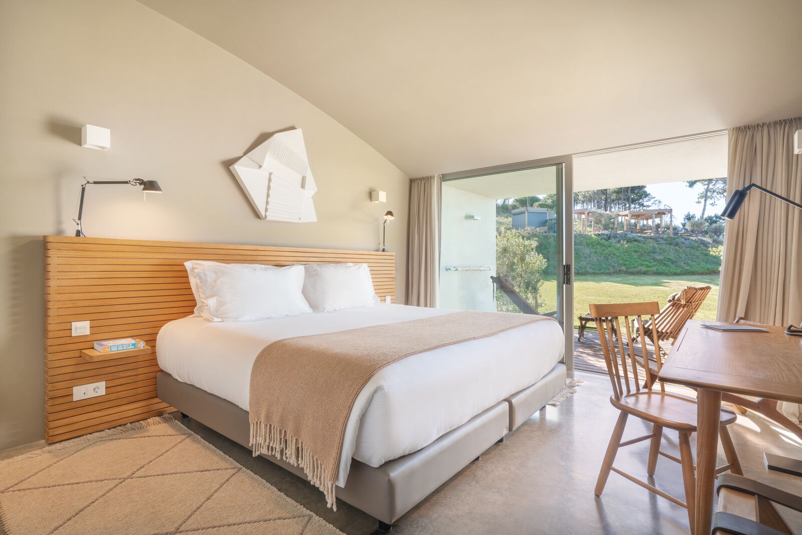 Quarto com uma cama grande, cabeceira de madeira, escrivaninha com cadeiras e vista para o exterior.
