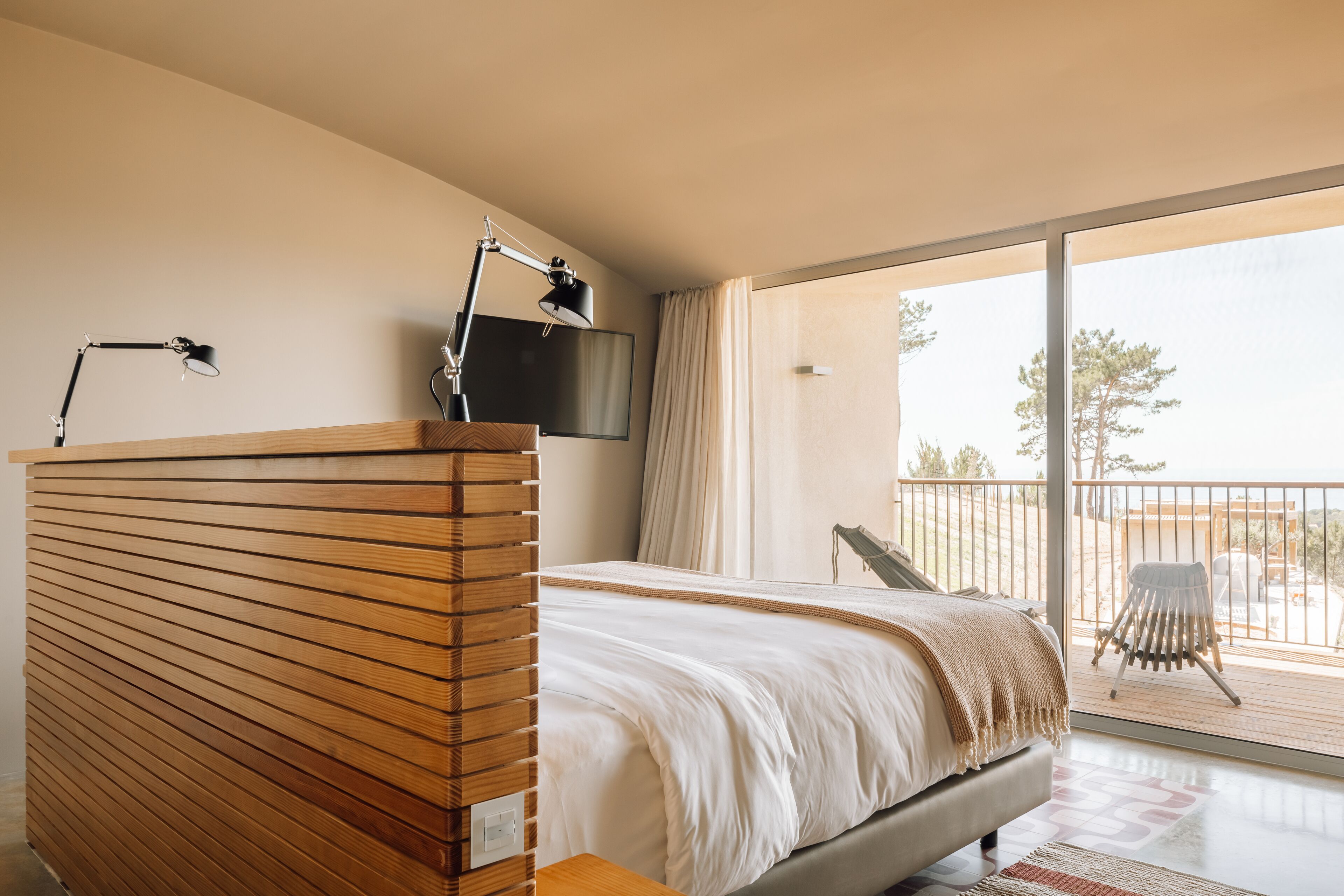 A bedroom with a wooden headboard, a bed with white bedding, a bedside table, a TV mounted on the wall, and a balcony with a chair.