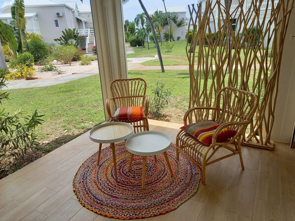 A patio with wicker chairs and a round table, overlooking a well-maintained garden and a white building in the background.