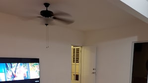 En stue med fjernsyn, ventilator, sofa med afdækning og et spisebord med stole.