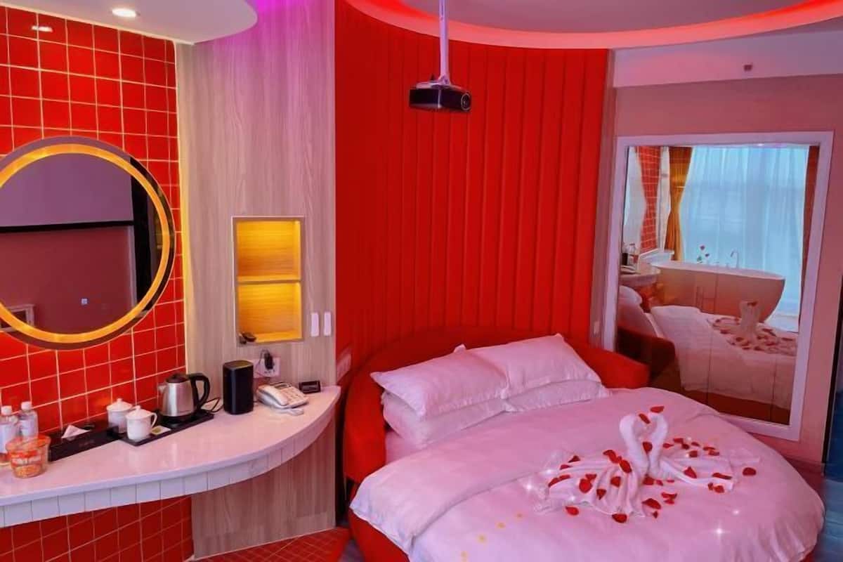 A hotel room with a red and white color scheme, a bed with rose petals, a bedside table with a kettle, a mirror, and a window with curtains.