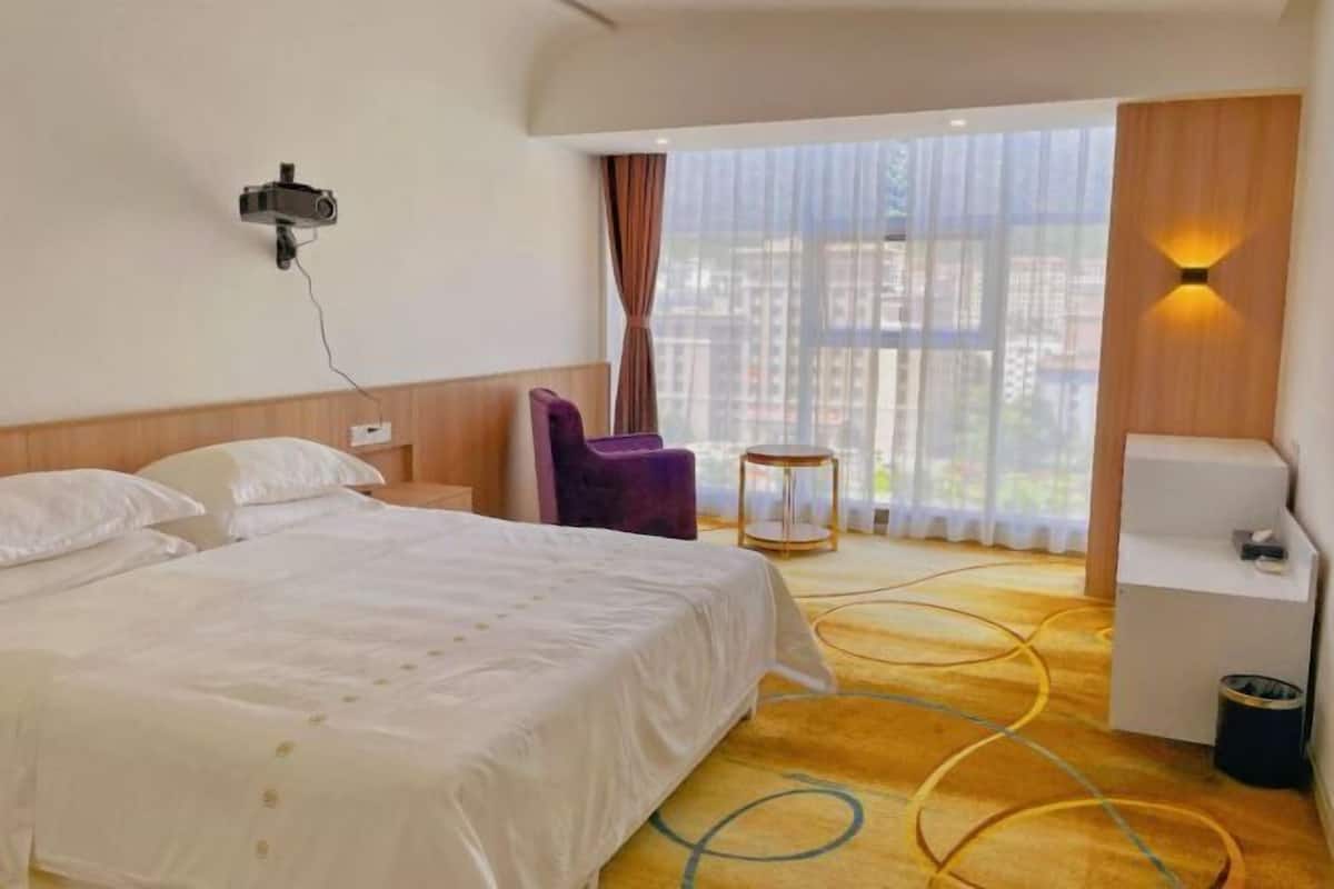 A hotel room with a bed, a chair, a small table, a wall-mounted projector, a window with curtains, and a view of buildings outside.