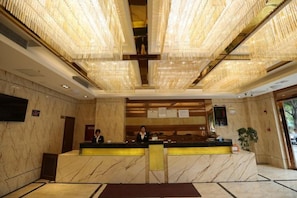 Un hall d’hôtel avec un comptoir de réception en marbre, un grand lustre et un téléviseur à écran plat fixé au mur.
