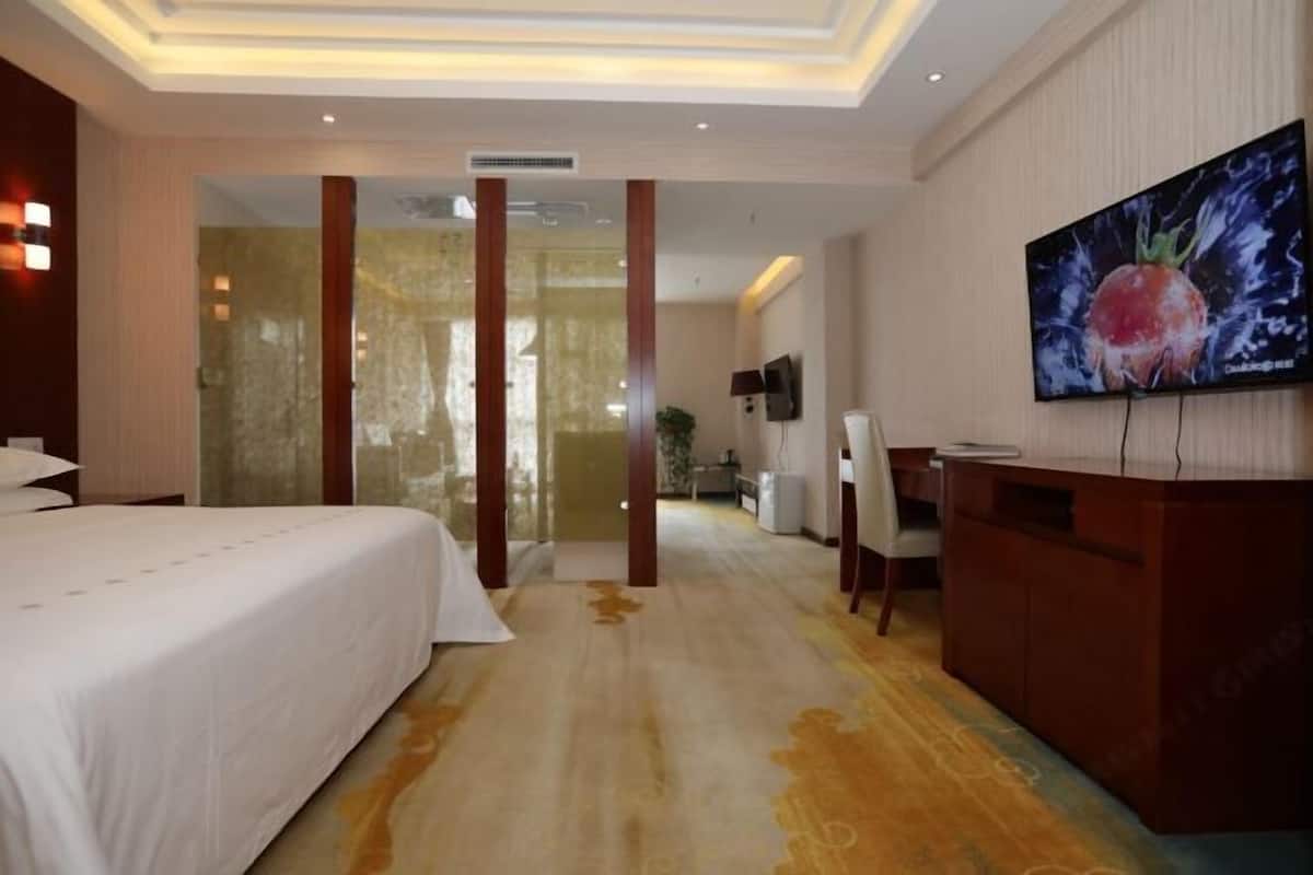 A hotel room with a large bed, a desk with a TV, and a glass partition.
