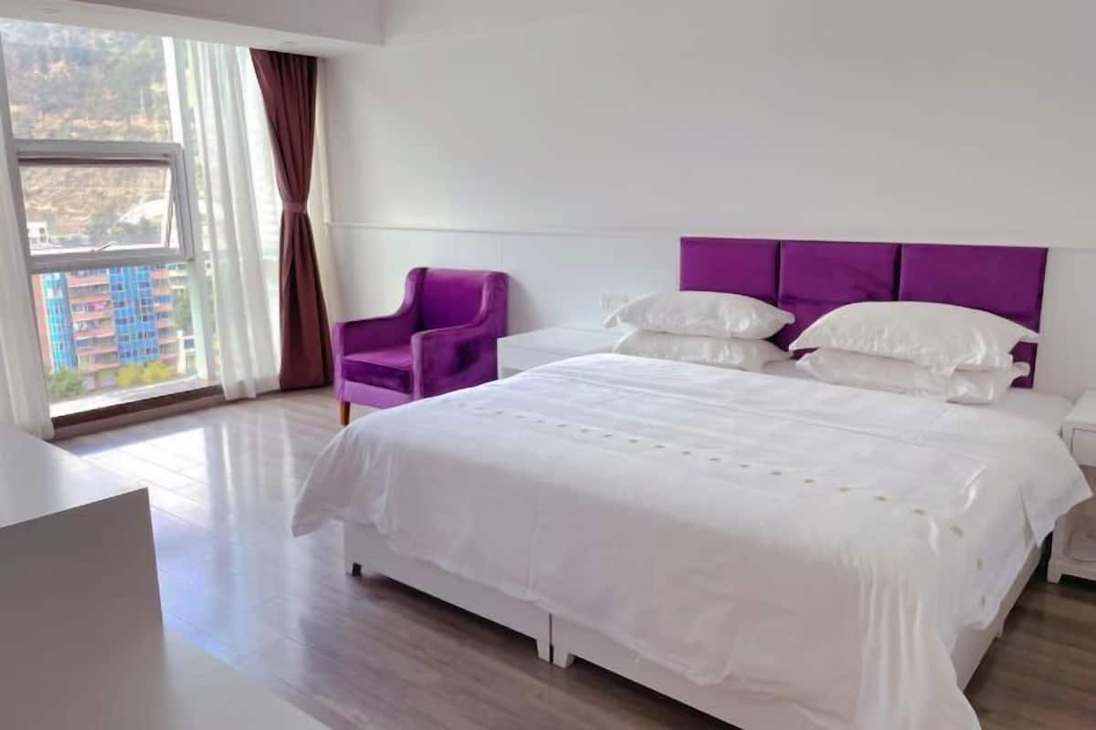 A modern hotel room with a large bed, a purple armchair, and a view of buildings through a window.