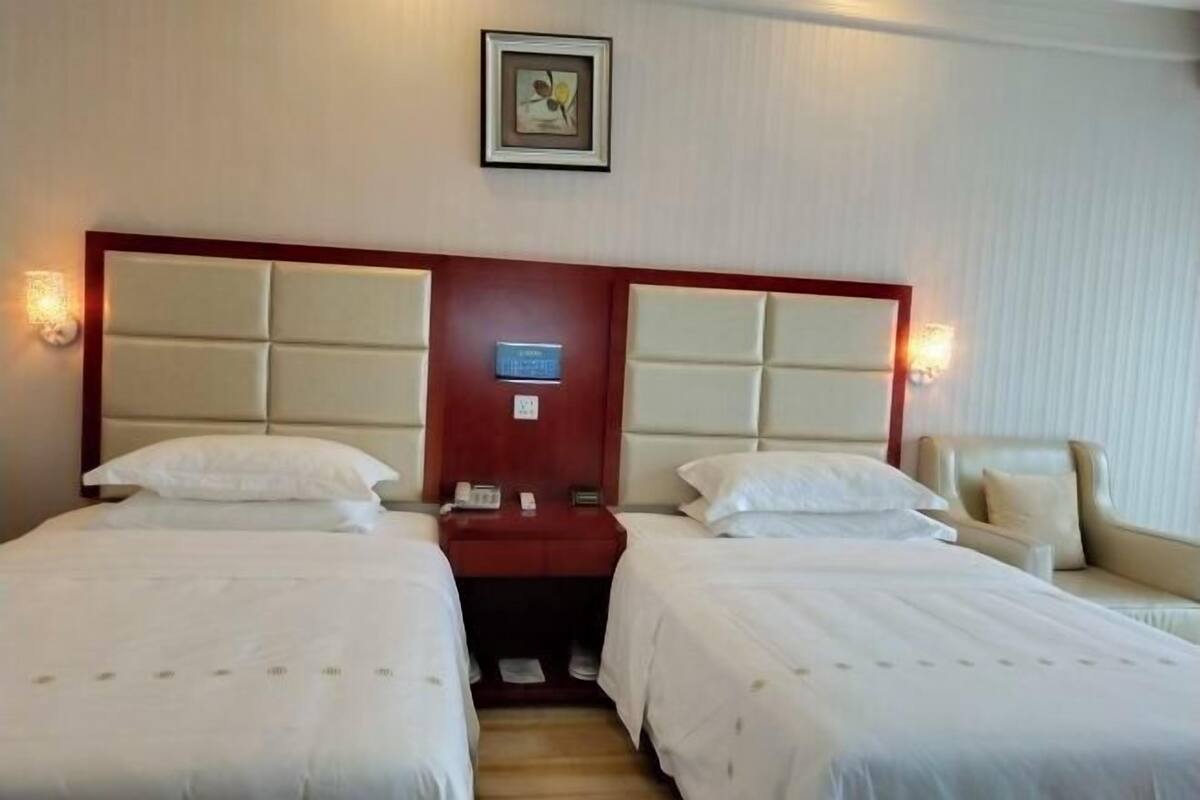 A hotel room with two beds, a wooden headboard, bedside tables with lamps, a small desk, and a framed picture on the wall.