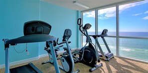 A gym with cardio machines and a treadmill, large windows with a view of the ocean.