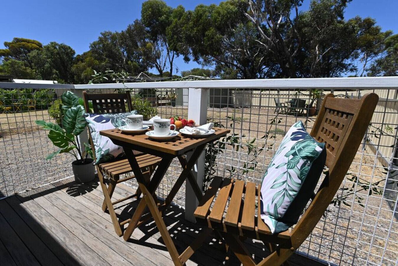 A wooden deck with a table set for two, a chair with a leaf-patterned cushion, and a potted plant.