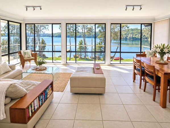 A spacious living room with large windows, a sofa, a coffee table, a bookshelf, and a dining area with chairs and a table.