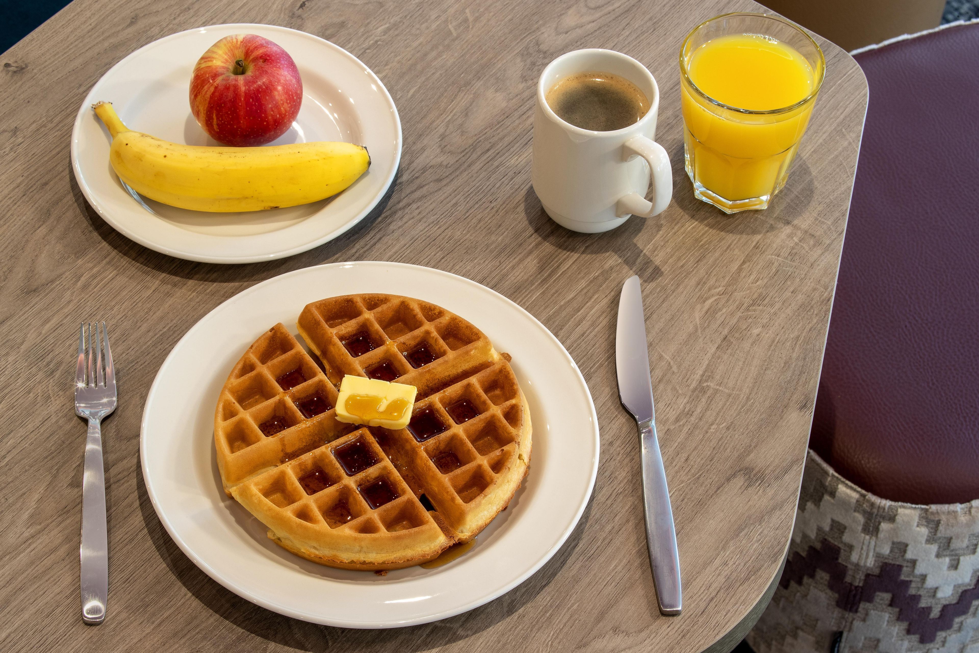 A breakfast setting with a waffle, apple, banana, coffee, and orange juice on a table.