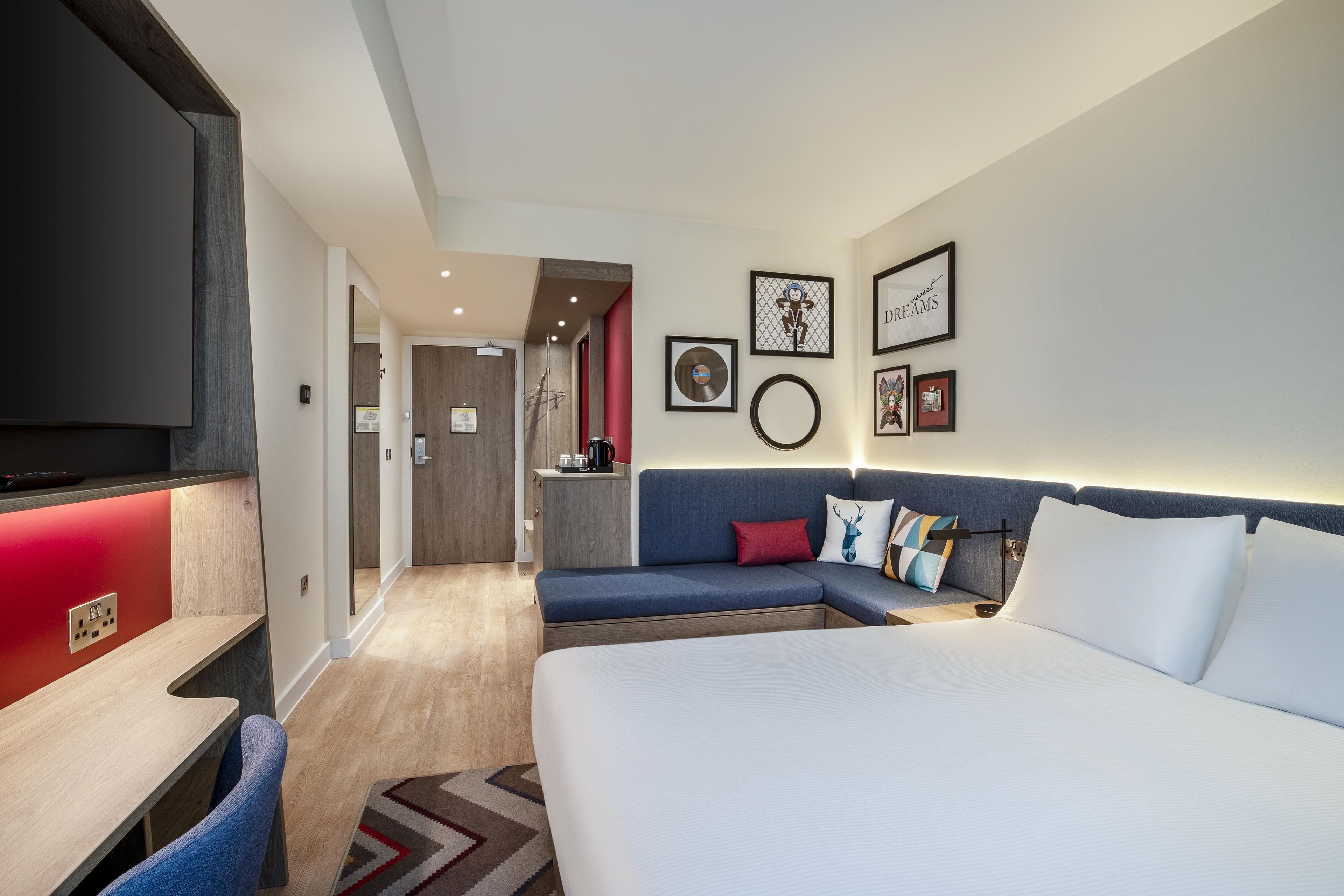 A hotel room with a large bed, a sofa, a desk, and a TV. There are framed pictures on the wall and a red accent wall.
