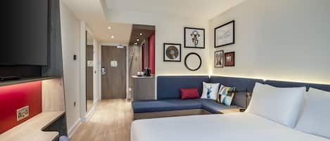 A hotel room with a large bed, a sofa, a desk, and a TV. There are framed pictures on the wall and a red accent wall.