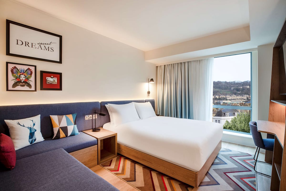 Habitación de hotel con cama, sofá, escritorio y un amplio ventanal con vista a la ciudad.