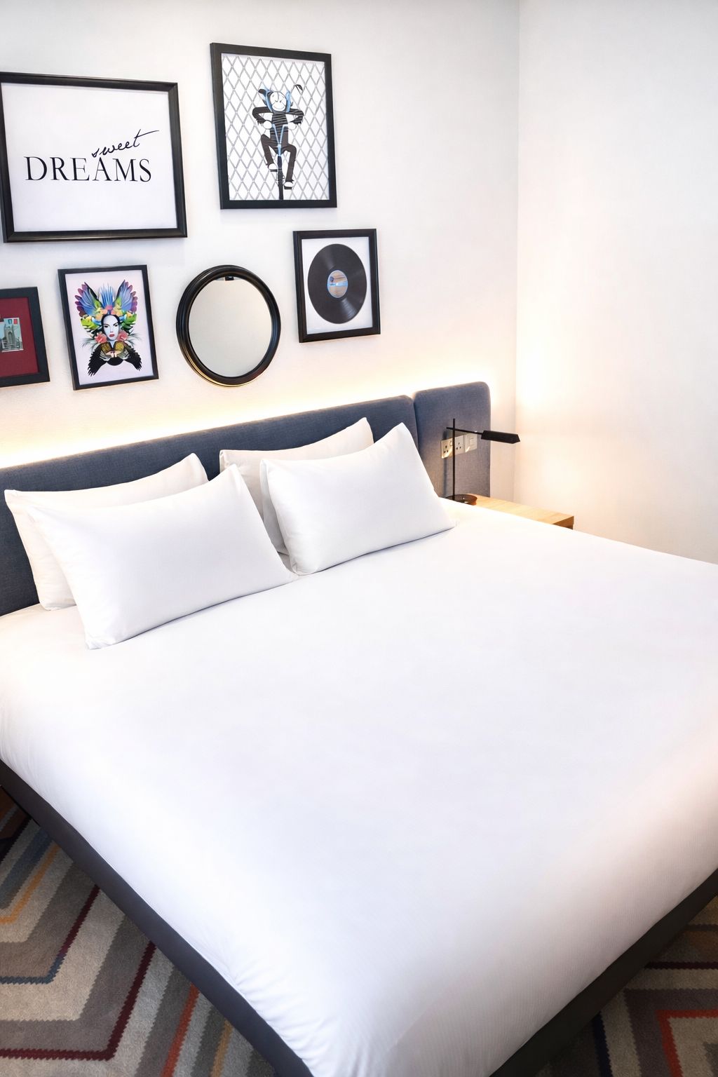 Una cama bien hecha con ropa blanca y dos almohadas, una pared con obras de arte enmarcadas incluyendo un disco de vinilo, y una mesita de noche moderna con una lámpara.
