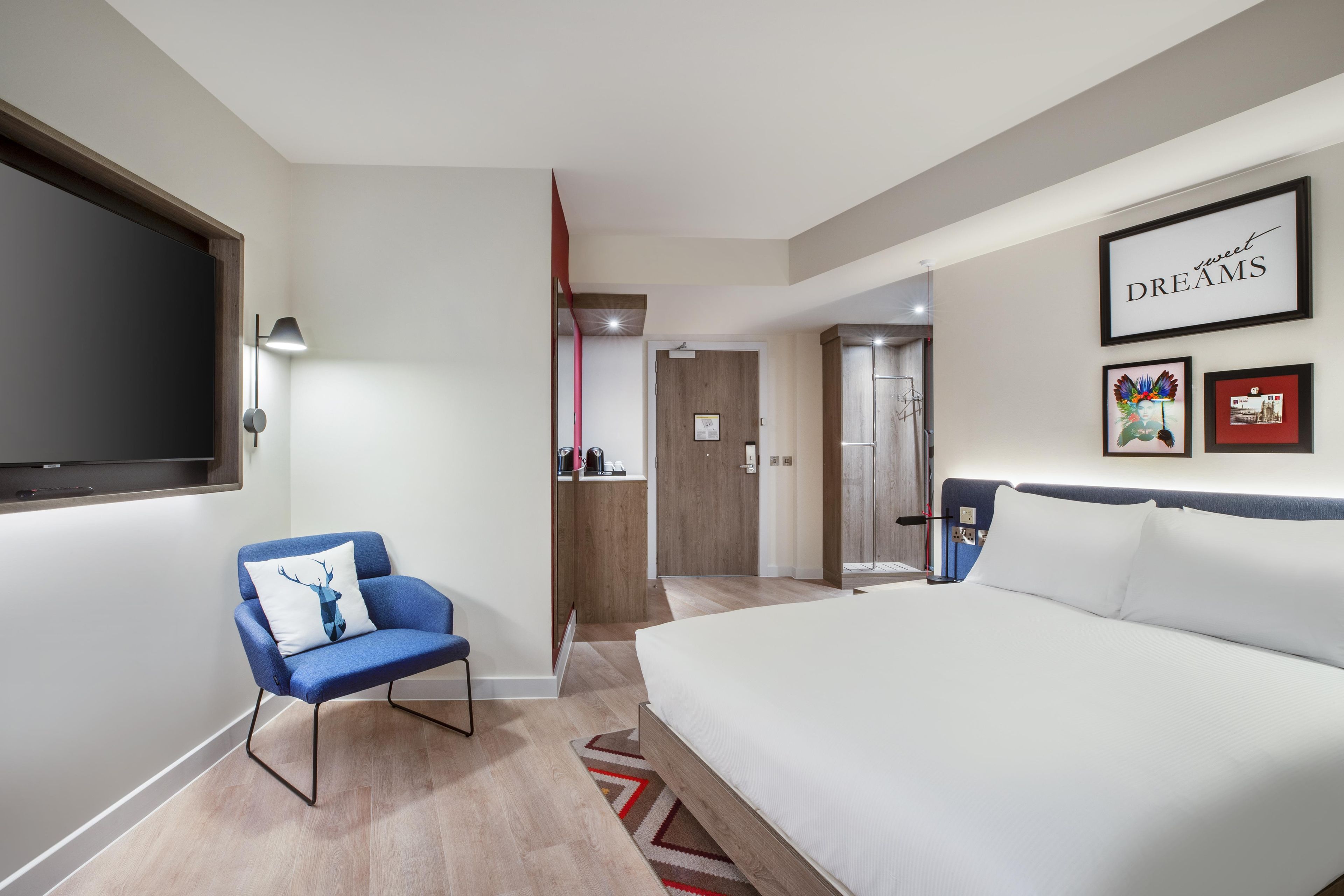 A modern hotel room with a large bed, a seating area with a blue chair, a flat-screen TV, and a framed artwork on the wall.