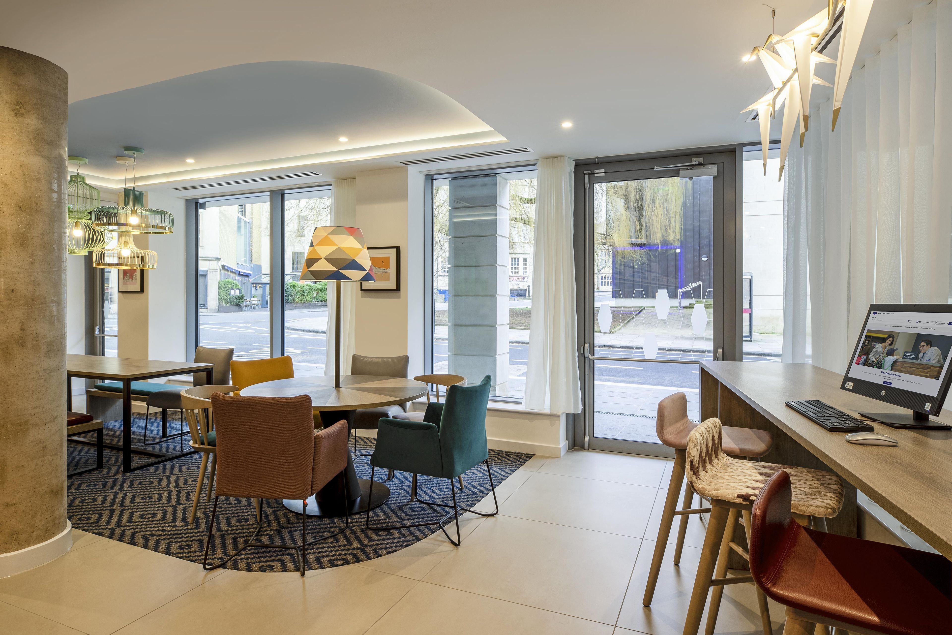 A modern hotel lobby with a seating area, a reception desk, and large windows offering a view of the street.