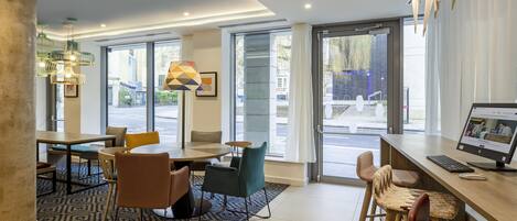 A modern hotel lobby with a seating area, a reception desk, and large windows offering a view of the street.