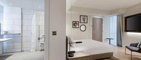 A modern hotel room with a bed, a TV, a small table, and a bathroom visible through an open door.