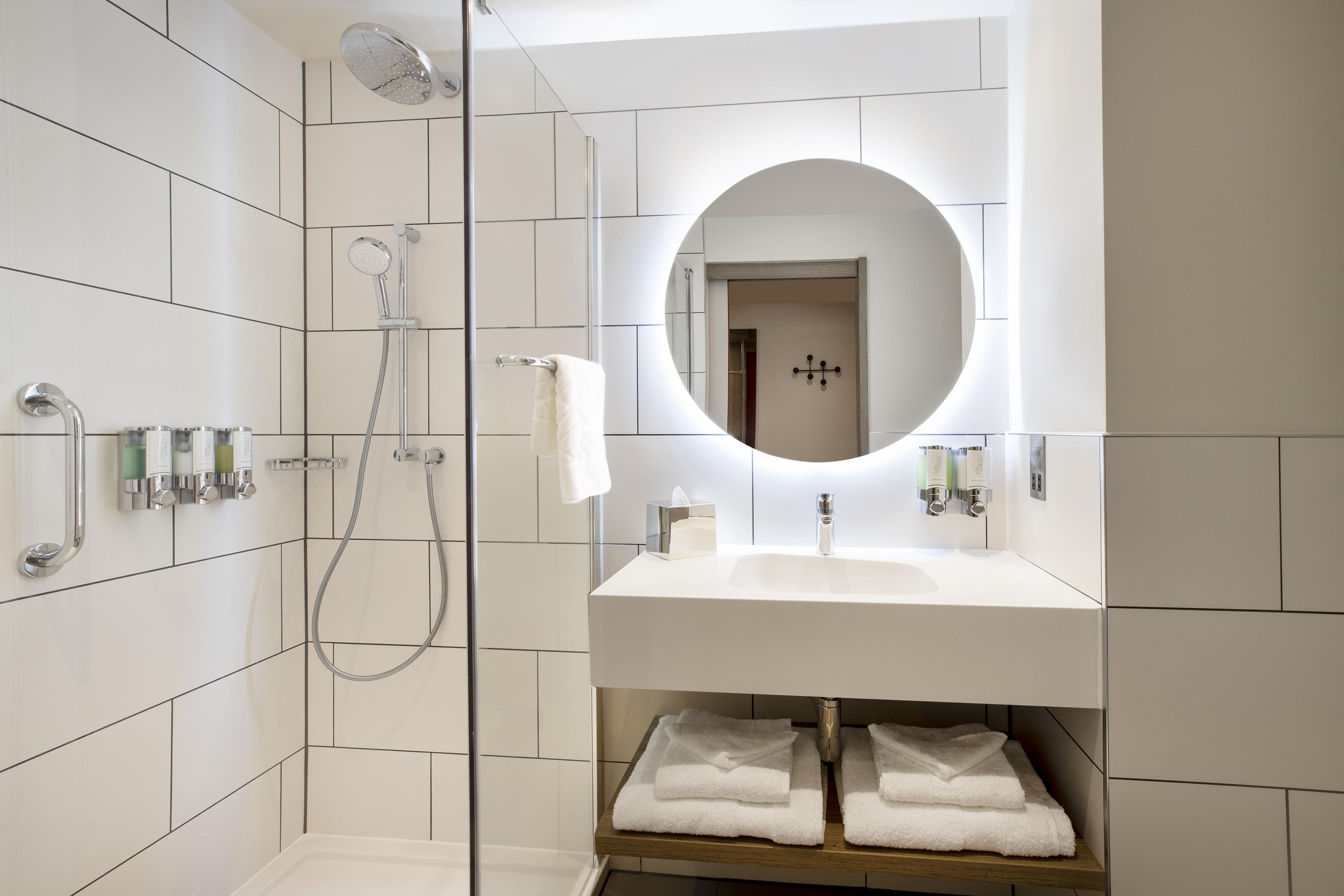 Ein modernes Badezimmer mit einer gläsernen Dusche, einem weißen Waschbecken mit Spiegel darüber und ordentlich gefalteten Handtüchern auf einem Regal.