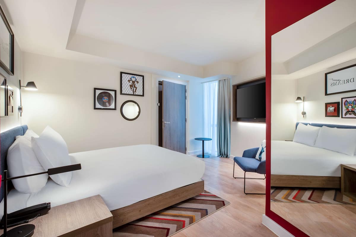 Una habitación de hotel moderna con una cama grande, un escritorio y una silla. Se ve una puerta que conduce a otra habitación y una ventana con cortinas.
