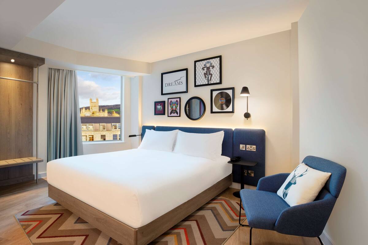 Una habitación de hotel moderna con una cama grande, un sillón azul y vistas a la ciudad.