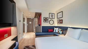 A modern hotel room with a large bed, a sofa, a desk, and a TV. There are framed pictures on the wall and a small kitchen area in the background.