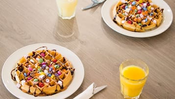 Two plates of waffles with colorful candies and chocolate syrup, a glass of orange juice, and a fork on a wooden table.