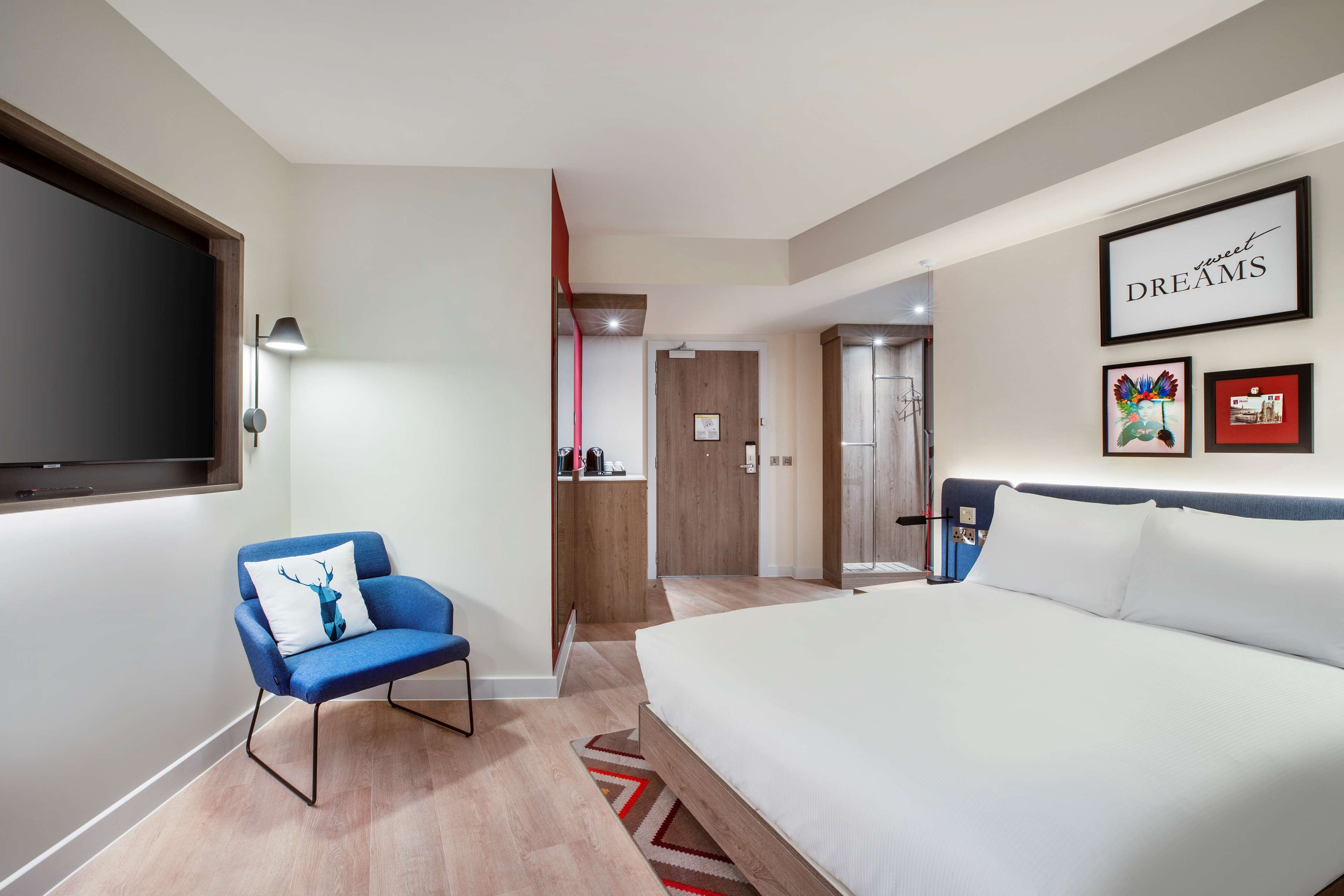 A modern hotel room with a large bed, a blue armchair with a deer pillow, a TV mounted on the wall, and a framed artwork with the words "sweet DREAMS".