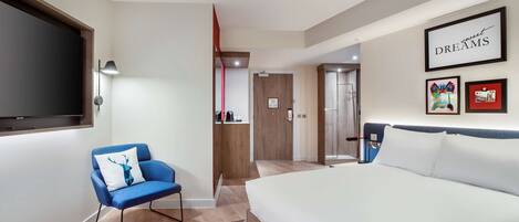 A modern hotel room with a large bed, a blue armchair with a deer pillow, a TV mounted on the wall, and a framed artwork with the words "sweet DREAMS".
