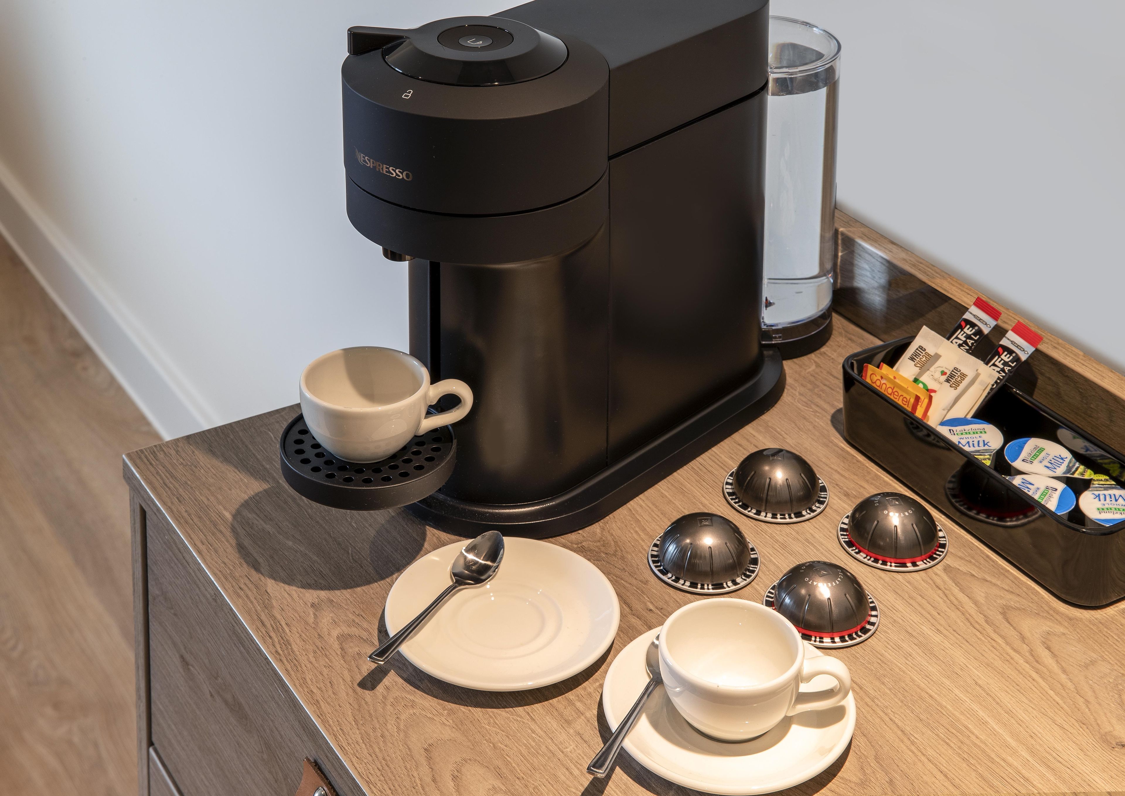 Una cafetera, una taza, un plato, una cuchara y un cartón de leche sobre una mesa de madera.