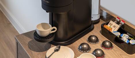 A coffee maker, a cup, a plate, a spoon, and a milk carton on a wooden table.