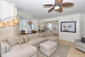 A living room with a sectional sofa, ottoman, ceiling fan, chandelier, dining area, kitchen, and a framed picture on the wall. - 10th Floor 3 Bedroom Condo at Silver Beach Towers (Destin)