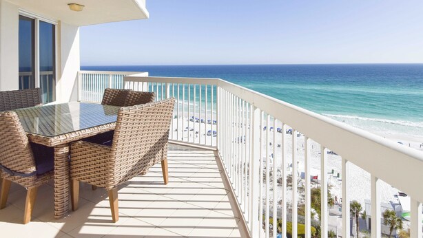 Ein Balkon mit einem Glastisch und Korbstühlen, der auf einen Strand mit weißem Sand und das Meer hinausblickt.