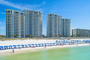 A beachfront hotel with multiple high-rise buildings, a sandy beach with blue umbrellas, and clear turquoise water.