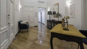 Un pasillo con piso de madera pulida, un sillón azul, una mesa con lámpara y un aparador con objetos decorativos.
