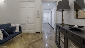 Un pasillo con un sofá azul oscuro, puertas blancas, una lámpara y un espejo sobre una consola de madera oscura.