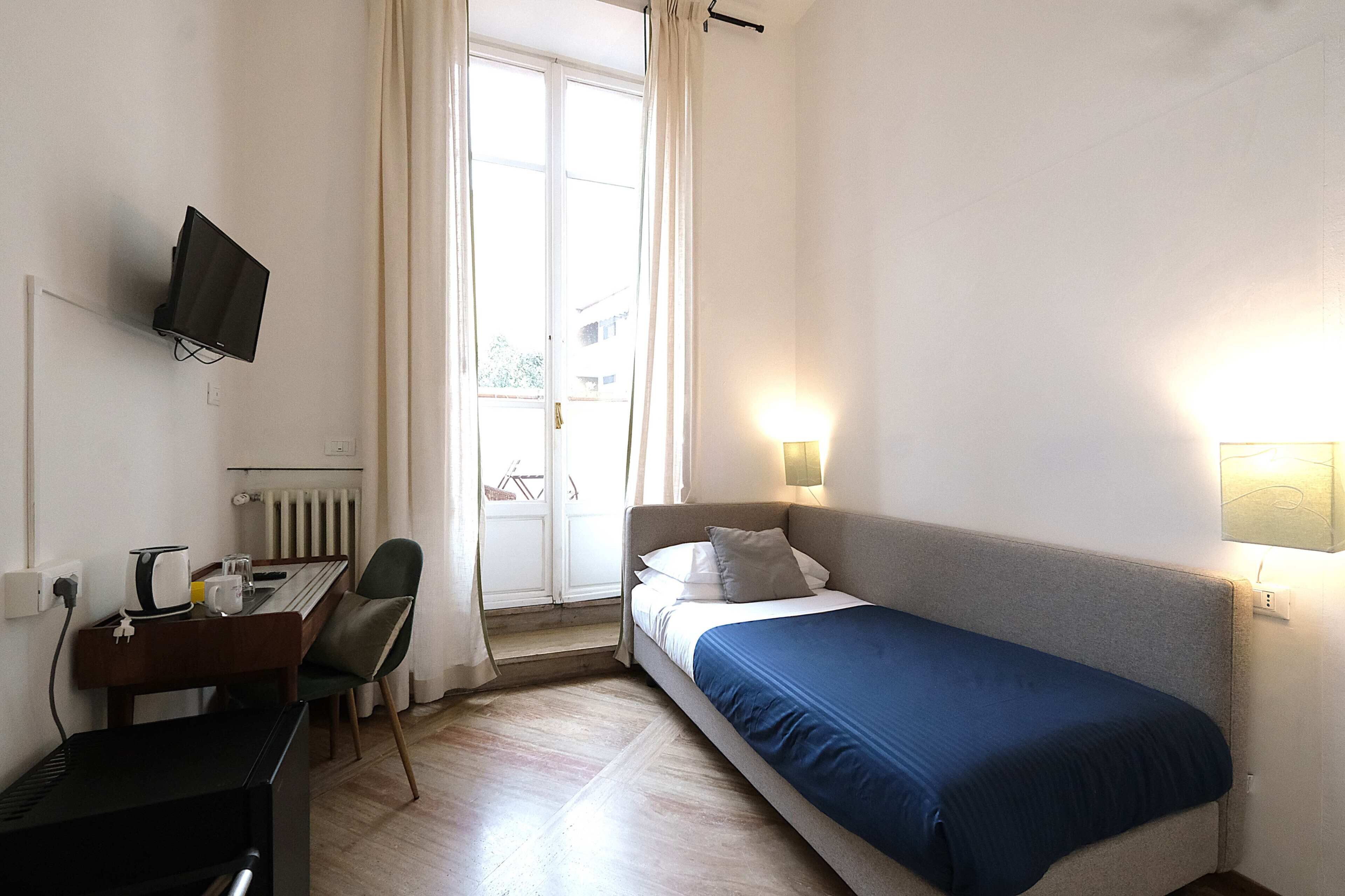 Quarto com cama, escrivaninha com computador, cadeira, televisão na parede e janela com cortinas.