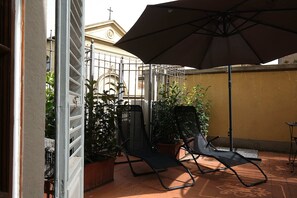 Eine Terrassenfläche mit einem großen Sonnenschirm, zwei Stühlen und Blumentöpfen.