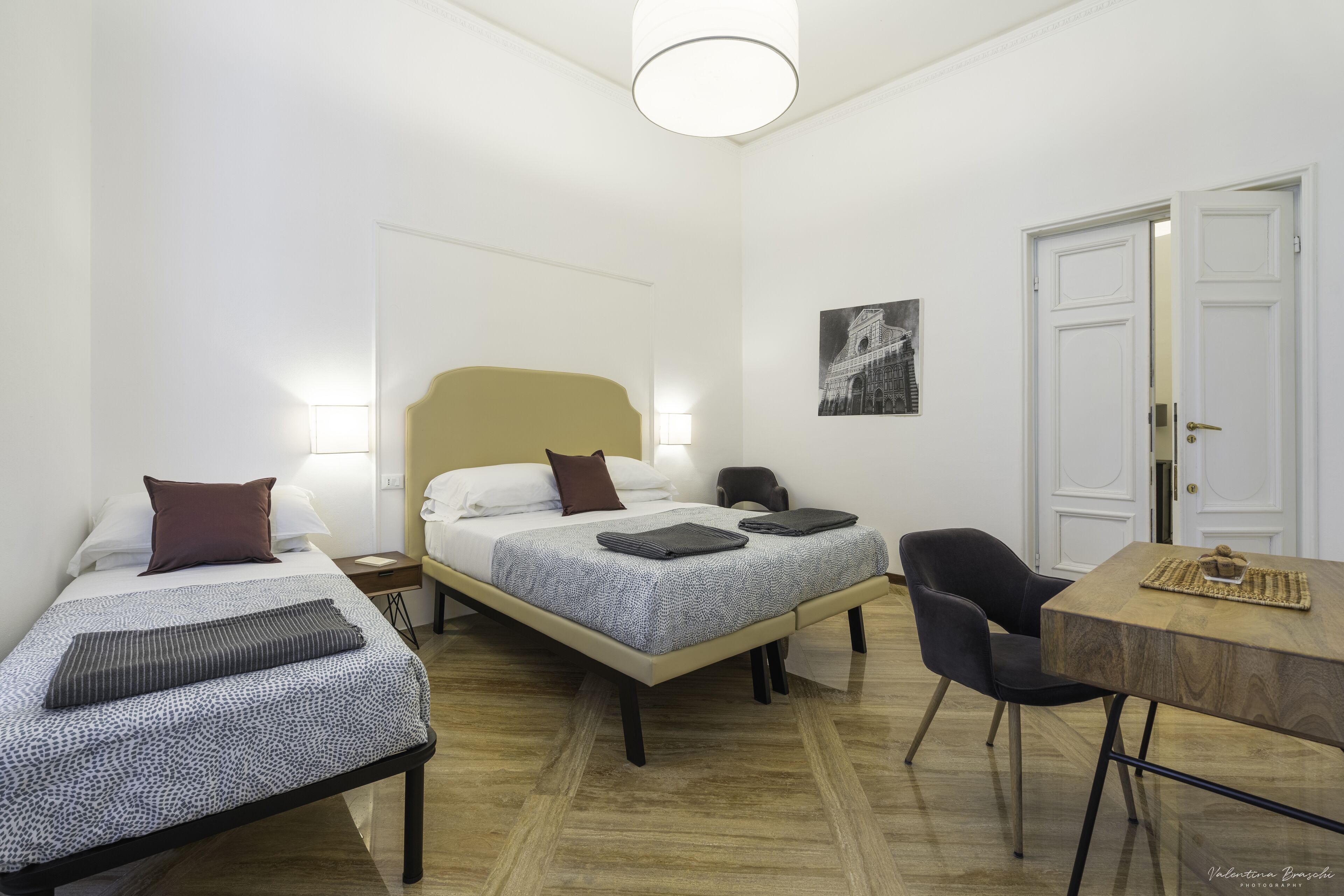 A hotel room with two single beds, a desk with a chair, and a door.